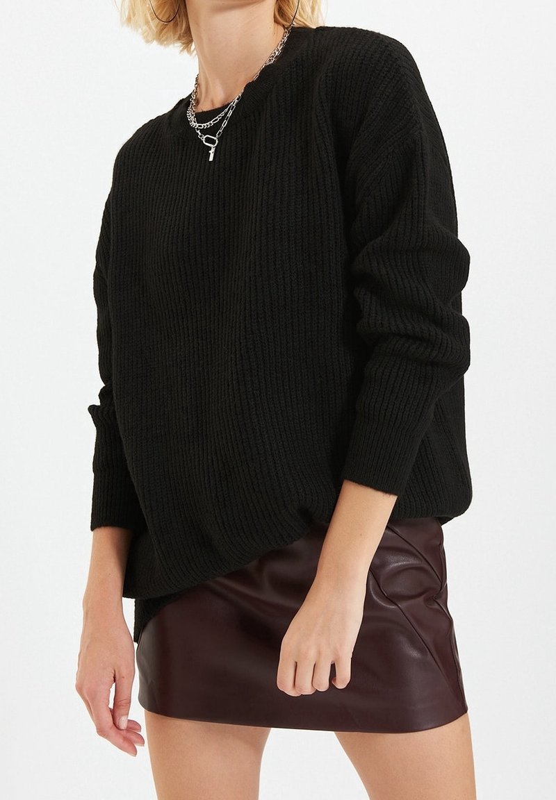 Femme portant un pull en grosse maille noir, un collier chaîne argenté et une mini-jupe en cuir marron foncé sur fond blanc.