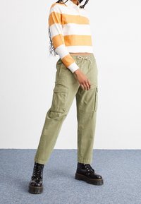 Orange-weiß gestreiftes langärmeliges Polohemd kombiniert mit hellolivgrünen Cargohosen mit Seitentaschen und Kordelzug in der Taille. Schwarze Schnürstiefel.