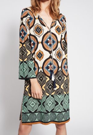 Long-sleeve knee-length dress with geometric diamond and circle patterns in cream, black, orange, teal, and blue, featuring a tassel tie neckline.