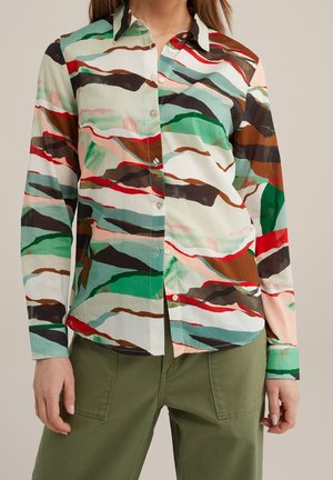Vrouw draagt een overhemd met lange mouwen met abstracte groene, bruine, rode, zwarte en witte patronen, gecombineerd met olijfgroene broek.