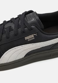 Sapatilha de couro preto com uma faixa em camurça cinza, sola de borracha texturizada preta, atacadores pretos e a marca PUMA em prata na lateral.