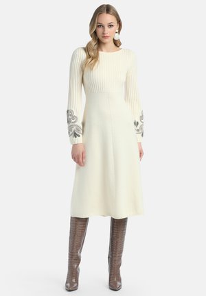 Ženska v smetnjakavi pleteni obleki z vzorčastimi rokavi, ki nosi velike biserne uhane in rjave krokodilne škornje do kolen, stoji.