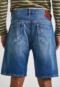 Pantalones cortos de mezclilla en azul claro con efecto desteñido. Presenta dos bolsillos traseros con detalles de costura y una etiqueta de cuero marrón en la cinturilla.