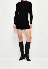 Woman wearing a black ribbed turtleneck sweater, black shorts with metallic buttons, and black knee-high heeled boots standing against a white wall.