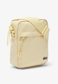 Kleine beigen Umhängetasche mit vorderer Reißverschlusstasche, verstellbarem Riemen und grünem Krokodillogo in der unteren rechten Ecke.