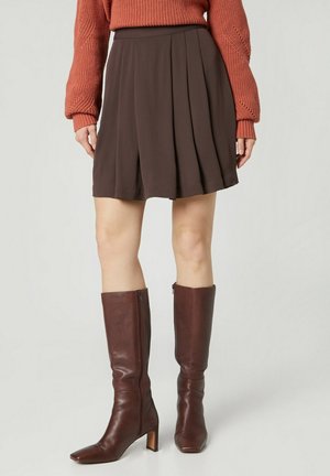Femme portant une jupe plissée marron, un pull en laine couleur rouille et des bottes en cuir brun foncé montantes jusqu’au genou avec des talons blocs.
