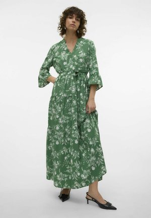 Robe maxi florale verte avec un décolleté en V, des manches trois-quarts et une taille nouée. Elle présente une jupe à volants et un tissu texturé. Chaussures à talons noires.