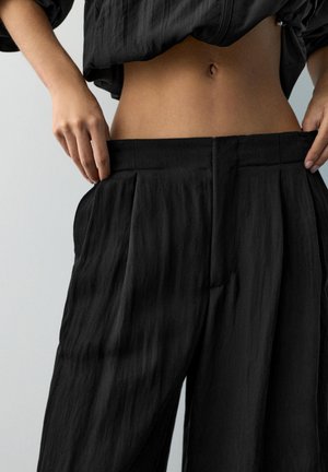 Persoană ține pantalonii negri largi la talie, dezvăluind abdomenul gol și o jachetă neagră scurtă asortată, cu fermoar.