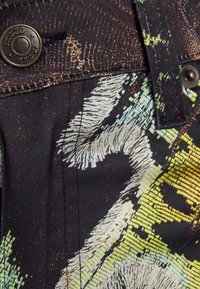 Close-up van een doek met een patroon in groen, geel en zwart. Het bevat een metalen knoop en zichtbare stikdetails. Aardse ontwerpelementen.