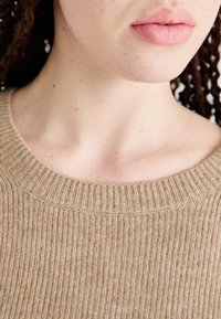 Suéter beige acanalado con cuello redondo y un acabado texturizado, que presenta puntadas visibles y un primer plano de piel sin adornos.