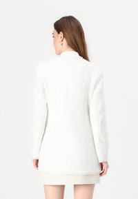 White tweed blazer with a textured surface, featuring a classic collar and long sleeves, seen from the back. Short length above a beige skirt.