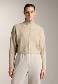 Beige gerippter, verkürzter Pullover mit hohem Kragen und halbem Reißverschluss, ausgestattet mit langen Ärmeln und einem strukturierten Strickmuster.