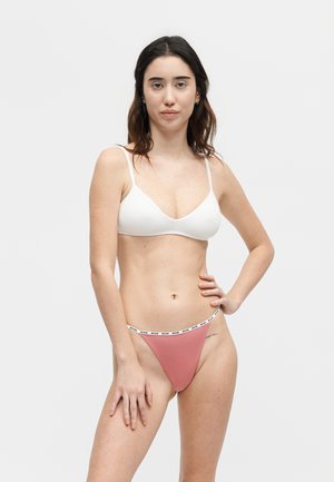 Donna in piedi con mano sul fianco che indossa reggiseno bianco e slip rosa a vita alta con fascia con logo su sfondo semplice.
