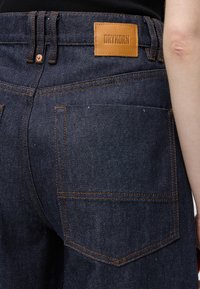 Gros plan sur la poche arrière d'un jean en denim bleu foncé avec des surpiqûres orange et un patch en cuir beige marqué Drykorn à la taille, à côté d'un bras nu.