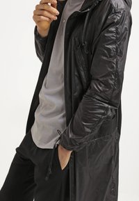 Manteau long noir, léger, avec capuche et poches zippées, présentant une texture lisse et brillante, avec des cordons de serrage ajustables. Porté sur un haut gris.