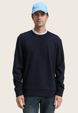 Marineblauwe sweatshirt met een ronde halslijn en lange mouwen, met ribgebreide manchetten en zoom. Draagt over een wit T-shirt. Lichtblauwe pet.