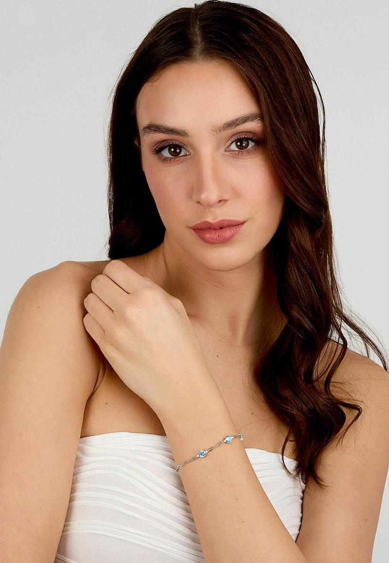 Braccialetto d'argento con tre pietre blu, caratterizzato da un design a catena delicata. Modella che lo indossa al polso su uno sfondo chiaro.