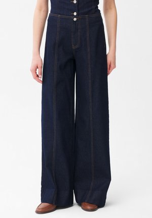 Donna che indossa pantaloni in denim blu scuro a vita alta e gamba larga con cuciture a contrasto e mocassini marroni, in piedi davanti a uno sfondo bianco.