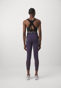 Sujeción deportiva negra con espalda con forma de llave, combinada con leggings morados que cuentan con detalles de bloqueo de color. La modelo lleva zapatillas ligeras.
