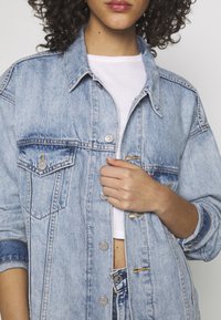 Denimová bunda v světle modré barvě s kovovými knoflíky, dvěma náprsními kapsami a volným střihem. Má kontrastní prošití a ohrnuté rukávy.