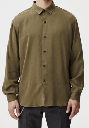 Homme portant une chemise boutonnée à manches longues couleur vert olive avec des boutons noirs et un pantalon sombre, les mains détendues le long du corps.