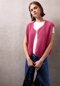 Fluffig rosa väst med knäppning fram, ihop med en vit t-shirt och blå jeans, hålls med en svart väska. Betongbakgrund.