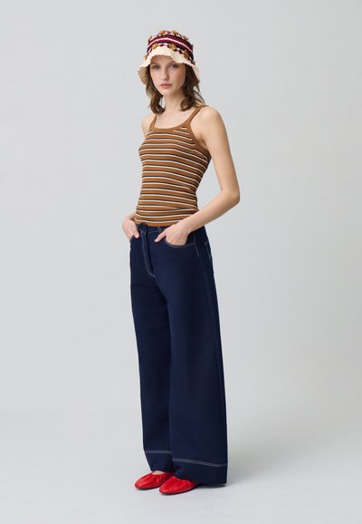 Débardeur rayé marron à côtes avec des bretelles fines, associé à un pantalon large bleu marine et des chaussures à enfiler rouges. Chapeau seau en tricot multicolore.