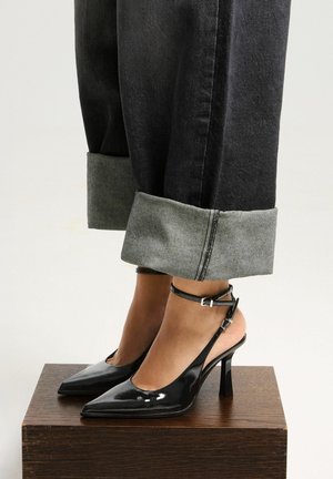 Black patent slingback heels with pointed toes and a thin ankle strap, paired with cuffed black jeans featuring a contrasting gray hem.