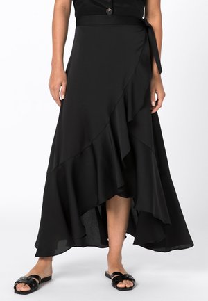 Femme portant une jupe portefeuille noire à ourlet asymétrique à volants et des sandales plates noires, debout devant un fond blanc.