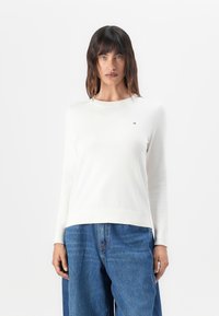 Hvit langermet genser med rund hals, laget av et glatt strikket stoff, med en liten logo på brystet og rett kant.