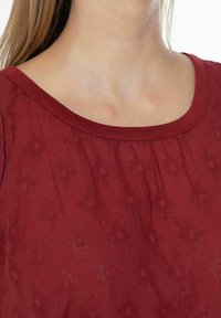 Top bordeaux con scollatura rotonda, tessuto testurizzato con motivi sottili e cuciture arricciate lungo la scollatura per un tocco di dettaglio in più.