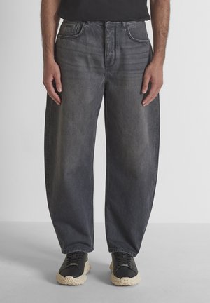 Uomo che indossa jeans neri slavati a vestibilità larga e sneakers nere con suola bianca, in piedi su un pavimento grigio chiaro davanti a uno sfondo semplice.