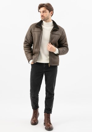 Man draagt een bruine shearling jas, een witte gebreide trui, zwarte broek en bruine leren laarzen, staand met één hand in de zak.