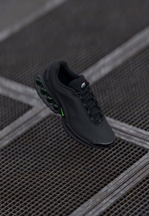 Chaussure de sport noire avec semelle coussin d'air verte et lacets, affichée au-dessus d'une surface métallique en grille.