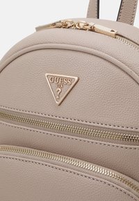 Sac à dos en simili cuir texturé beige avec deux compartiments zippés dorés et une plaque logo triangulaire dorée à l'avant.