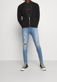 Cárdigan negro de punto con cremallera sobre una camiseta negra con texto naranja, combinado con jeans ajustados rasgados de un azul claro y zapatillas negras de caña alta.