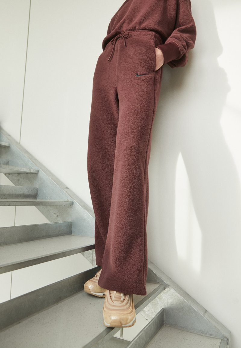 Pantalons de survêtement en polaire bordeaux avec une coupe décontractée, taille à cordon et une poche latérale avec un petit logo noir. Portés avec des baskets beiges.