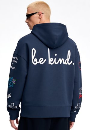 Uomo con capelli corti e biondi indossa una felpa con cappuccio blu navy con testo bianco "be kind." sulla schiena e varie piccole grafiche colorate sulle maniche.