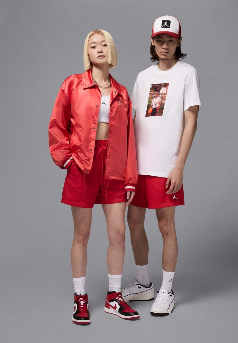 Rote Satin-Bomberjacke, rote Shorts, weißes Crop-Top und rot-schwarze Sneakers. Männliche Person in weißem T-Shirt mit Grafik, rot-weißem Cap. einfacher Hintergrund.