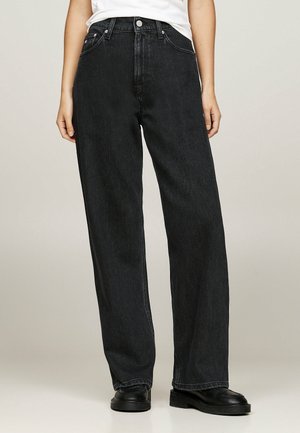 Persoon draagt losse zwarte jeans en zwarte schoenen, staat tegen een neutrale achtergrond met ontspannen handen langs het lichaam.