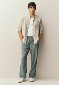 Camicia beige leggera di lino con bottoni sopra una t-shirt bianca, abbinata a pantaloni larghi azzurri chiari e scarpe da ginnastica bianche, mani nelle tasche.