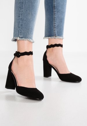 Jambes portant un jean bleu à ourlet effiloché et des chaussures noires en daim à talons blocs avec des brides ondulées à la cheville et des boucles rondes sur un sol blanc.
