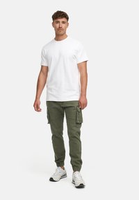 Homme debout portant un t-shirt blanc à manches courtes, un pantalon cargo vert olive et des baskets blanches, sur un fond clair uni.