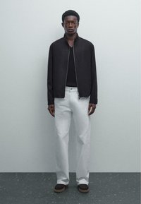 Young man standing against plain wall, wearing black zip-up jacket, black shirt, white pants, and dark brown shoes.