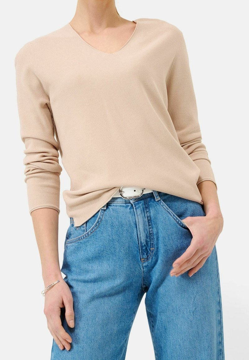 Personne portant un pull beige à col en V avec manches longues et un jean bleu taille haute avec une ceinture blanche, main dans la poche.