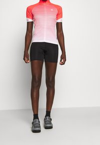 Koszulka rowerowa z krótkim rękawem w gradiencie różowym do białego, w połączeniu z czarnymi spodenkami. Posiada wysoki kołnierz i dopasowany krój. Nosi szare buty rowerowe.