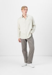 Hellbeige, langärmliges Hemd mit Knopfleiste, einer Tasche und dezenten vertikalen Streifen; kombiniert mit grauen Cordhosen und weißen Sneakers.
