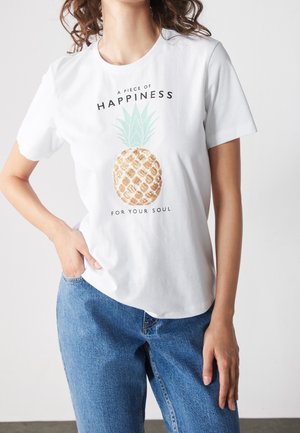 Kvinde iført hvid t-shirt med palietter i form af en ananas og teksten "Et stykke lykke til din sjæl," kombineret med blå jeans.