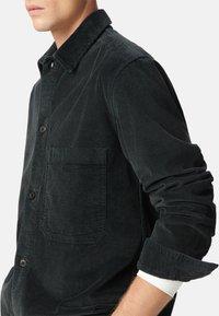 Giacca in corduroy nera con chiusura a bottoni, taschino sul petto e tessuto testurizzato. Presenta un colletto classico e maniche arrotolate.