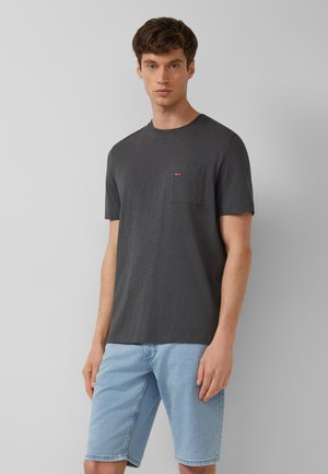 Młody mężczyzna stoi, ubrany w ciemnoszarą koszulkę z krótkim rękawem i kieszonką na piersi oraz jasnoniebieskie szorty jeansowe na tle jednolitej szarej ściany.